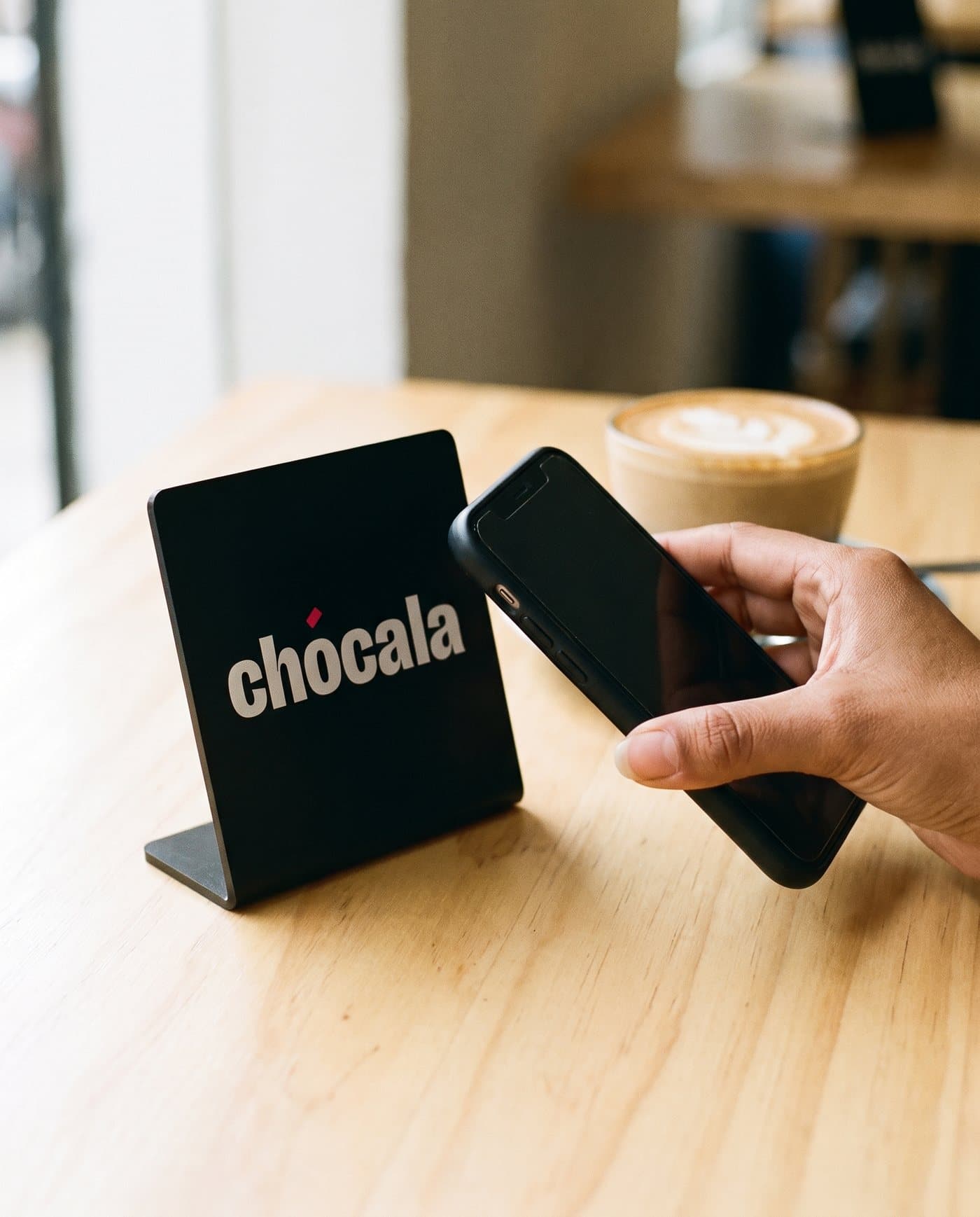 Soporte de Chócala en la mesa de una cafetería con una mano acercando un iPhone
